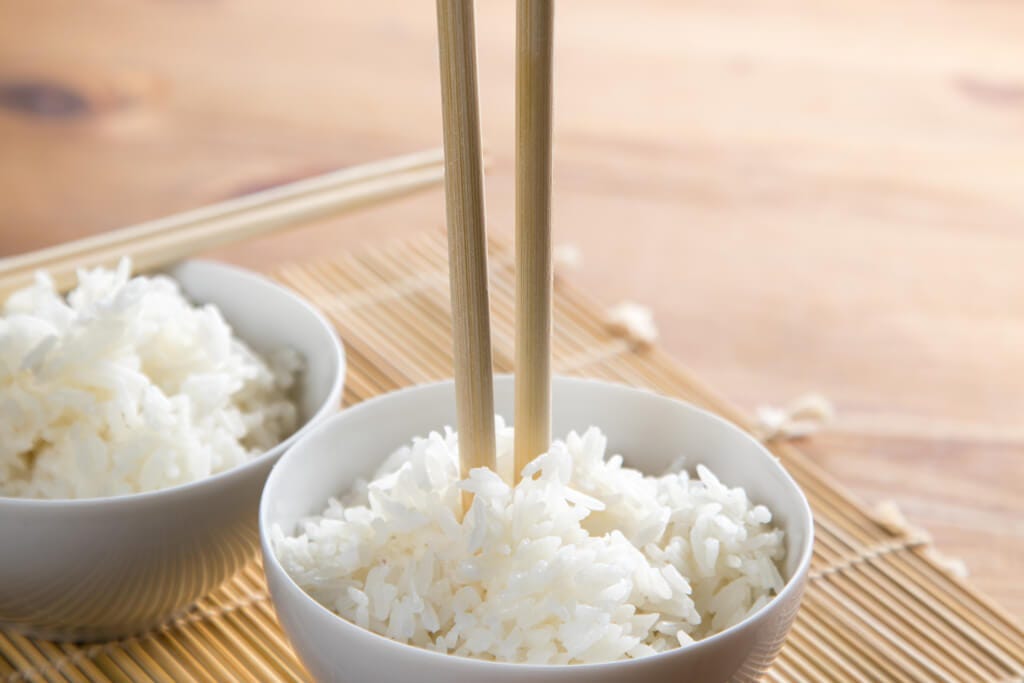 Two bowls of rice on a bamboo placemat on a wooden table with one bowl having a pair of chopsticks sticking upright in them.