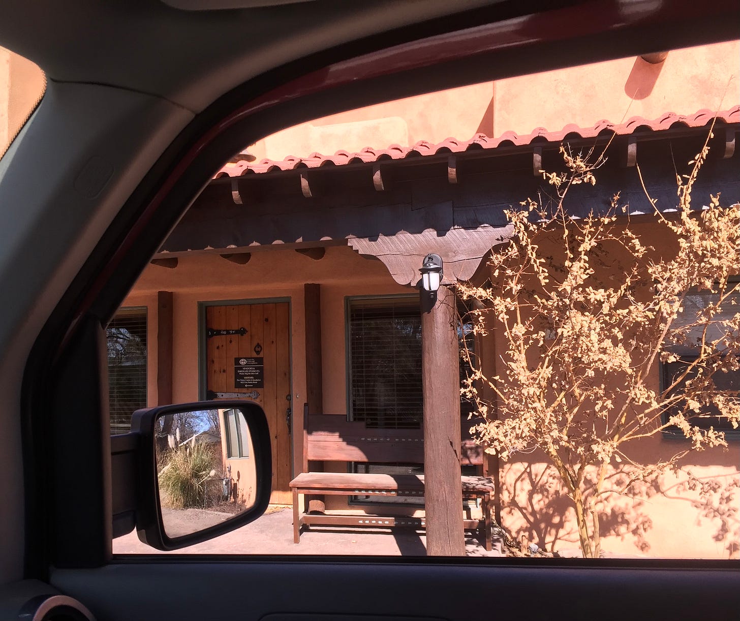 An adobe-style building with a wooden front door as seen through the open window of a car.