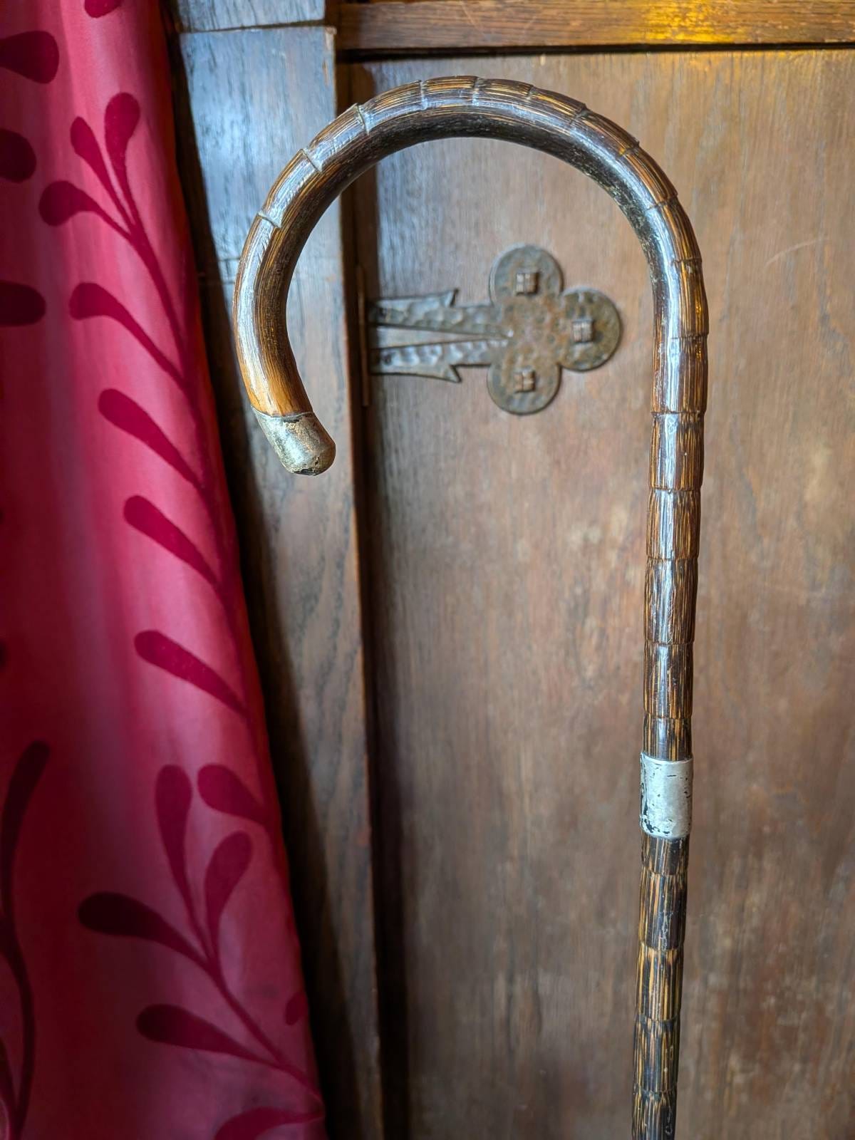 An antique-style wooden walking cane with a curved handle and metal fittings, standing against a wooden cupboard beside a red patterned curtain. An antique-style wooden walking cane with a curved handle and metal fittings, standing against a wooden cupboard beside a red patterned curtain.