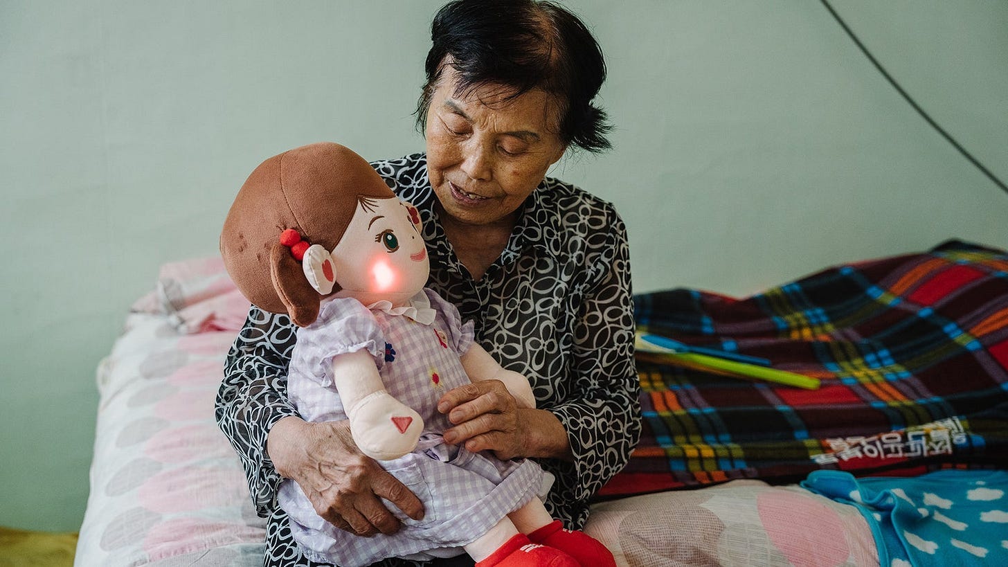 An elderly woman in a patterned blouse holds a soft doll with a light on its face, sitting on a bed covered in a pastel-colored sheet and various blankets.