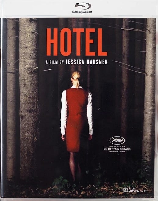 Blu-ray slipcover for Jessica Hausner’s Hotel (2004), featuring a woman in a red dress with her back turned, standing alone in a dark forest. The film title appears in bold red text against the trees.