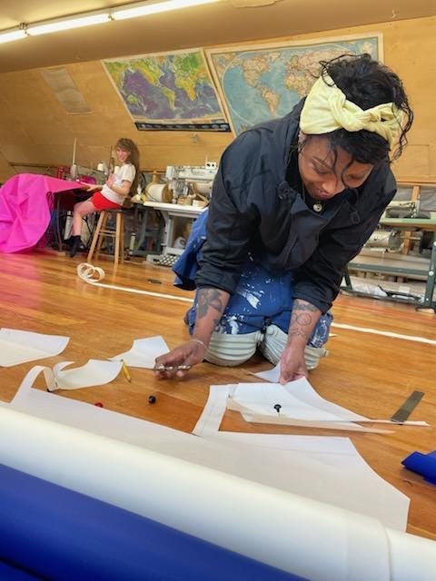 Two smiling artists in workshop. One sits at sewing machine, the other kneels over cut fabric on floor.