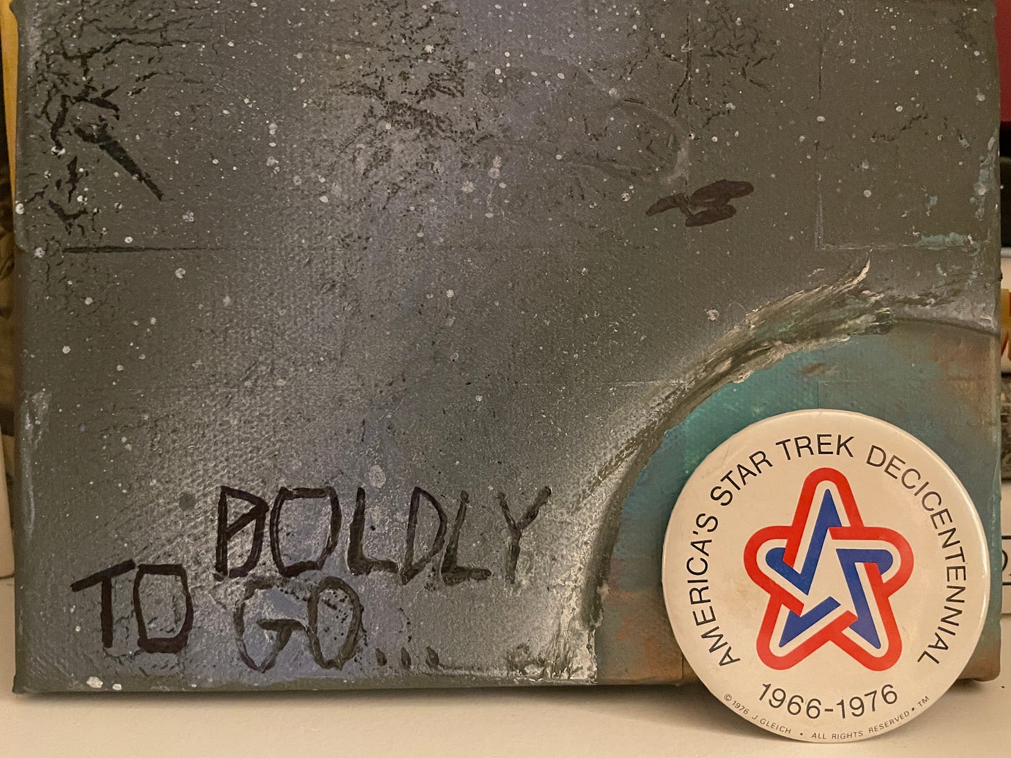 My Star Trek Decicentennial pin photographed in front of a painting my son did for me - the Enterprise orbiting a planet, and the words TO BOLDLY GO... painted on top