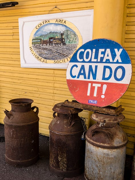 Three photos of Colfax. The first is the exterior of the local historical society building, an old wooden building painted yellow with large eaves and simple wooden brackets holding up the eaves. A door on the left has a hand written sign, "SHUT". From left to right is a bronze historical marker, a flag, an artistic lifesize cutout of a train conductor looking at his watch, an old clock with iron filligree, a wood burning stove, and a life-sized artist sculpture of a sunflower in a pot. The second photo are the rusty iron wheels and apparatus of an old rail car, including the armature for applying the brakes and a small section of rail to sit upon. Third photo is another exterior shot of the historical society, a closeup of objects outside the yellow building, including a primitive hand-painted sign of an old steam train on the tracks with mountains in the background set within a roundel around which the words, "COLFAX AREA HISTORICAL SOCIETEY" are painted. In front are some rusty milk barrels. A large round sign (bars of blue, white, and red with the words, "COLFAX CAN DO IT!" painted on it) sit on two of the barrels. A smaller sticker "TRUE" is affixed at the bottom of the sign.