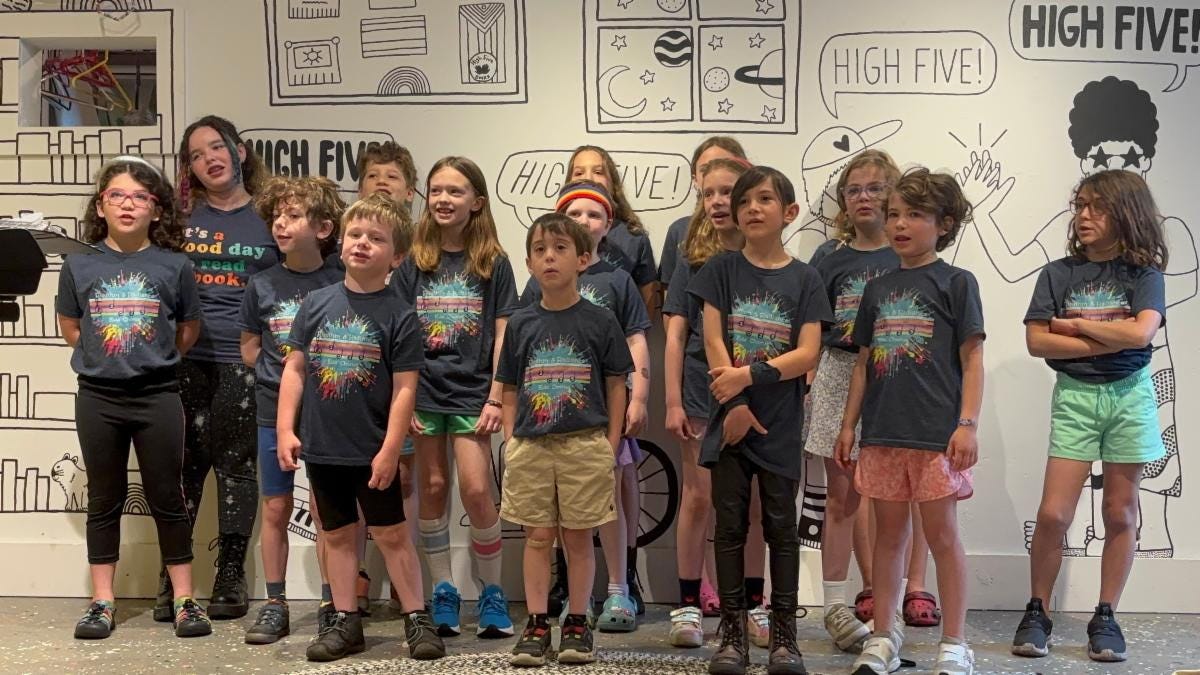 A bunch of kiddos in chorus tshirts stand in front of the High Five Books mural and sing