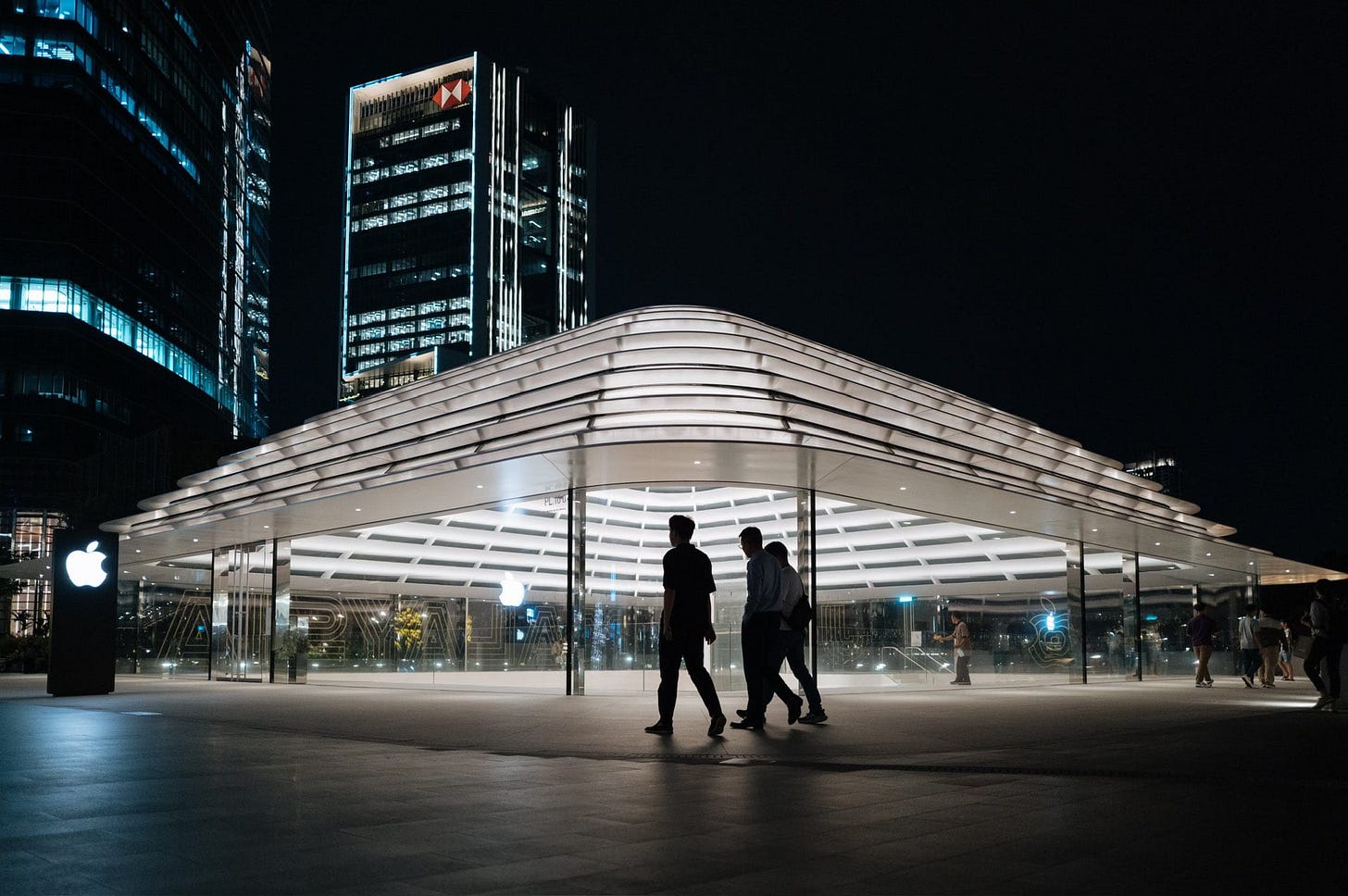 Apple The Exchange TRX at night, viewed from the park level. Three silhouetted figures walk past the store, which is brightly lit. Apple The Exchange TRX at night, viewed from the park level. Three silhouetted figures walk past the store, which is brightly lit.