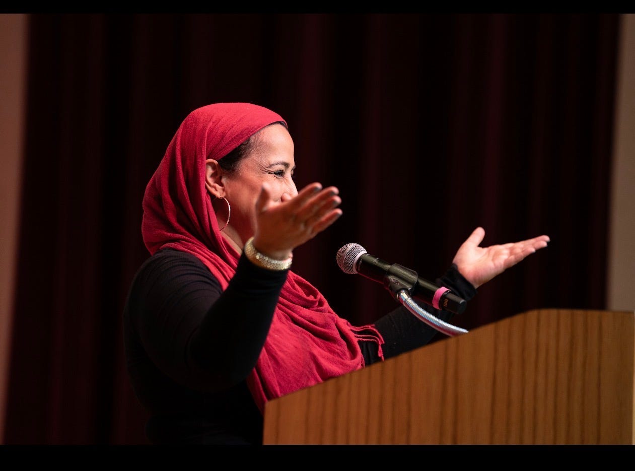 Najeeba Syeed, wearing a red hijab and hoop earrings, smiles at a microphone and gestures to the audience