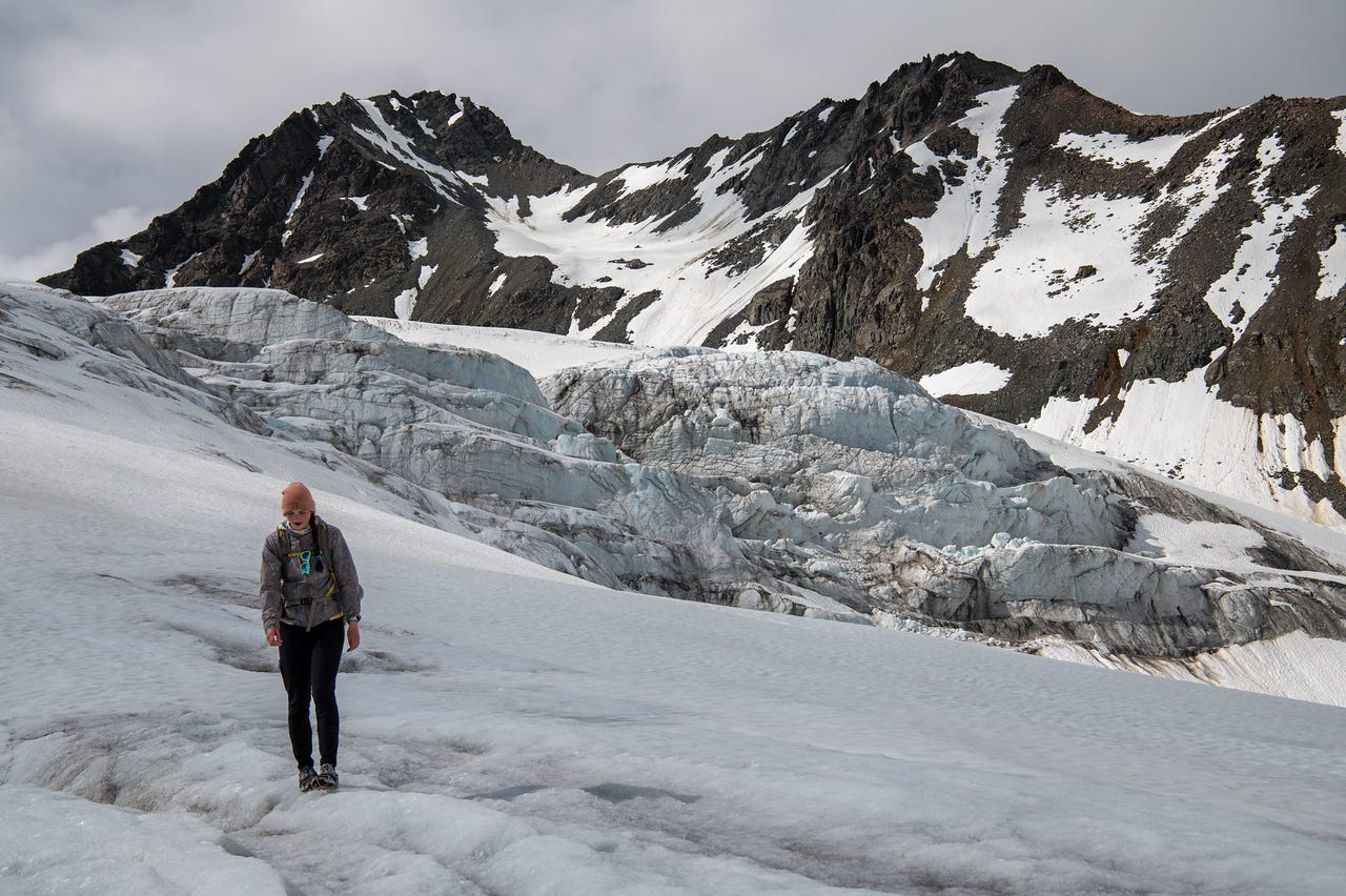 A girl in a peach beanie, grey patterned hoody, and black leggings walks toward the camera across bare white glacier ice, watching her footing. Behind her, the glacier steps up in long fractured tiers of grey-blue ice toward a saddle between two dark, snow-patched peaks under heavy overcast.