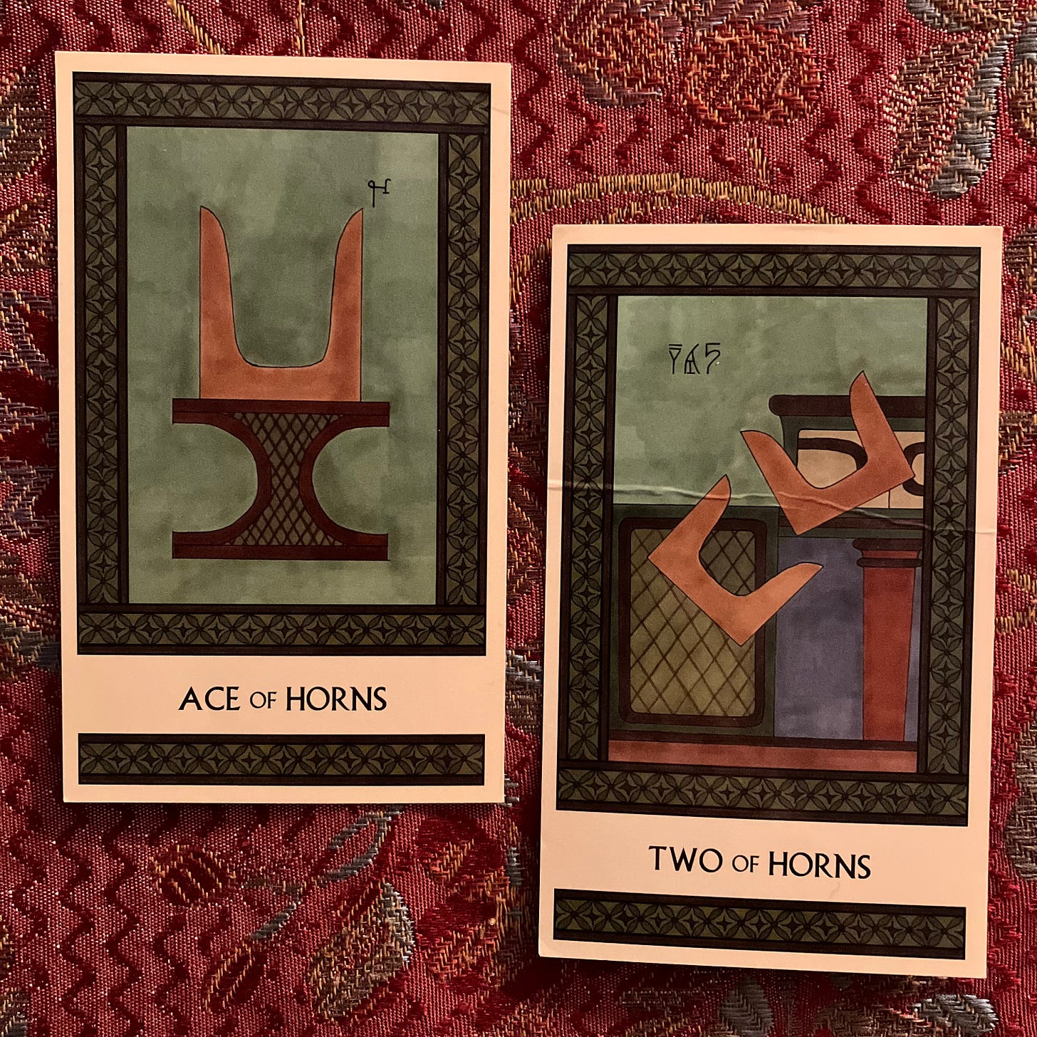 Two Minoan Tarot cards side by side on a red jacquard fabric background. Both cards are in shades of green and brown. The Ace of Horns shows a large pair of sacred horns atop an incurved altar. The Two of Horns shows two pairs of sacred horns tumbling off a shrine. 