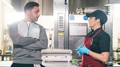 A man and woman stand in the school kitchen having a tense discussion