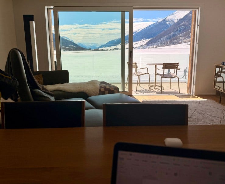 View from a wooden table with a laptop open on it, of a livingroom with a sliding door open onto a balcony with a bistro table and two chairs facing a snow-covered alpine valley on a bright, sunny day.