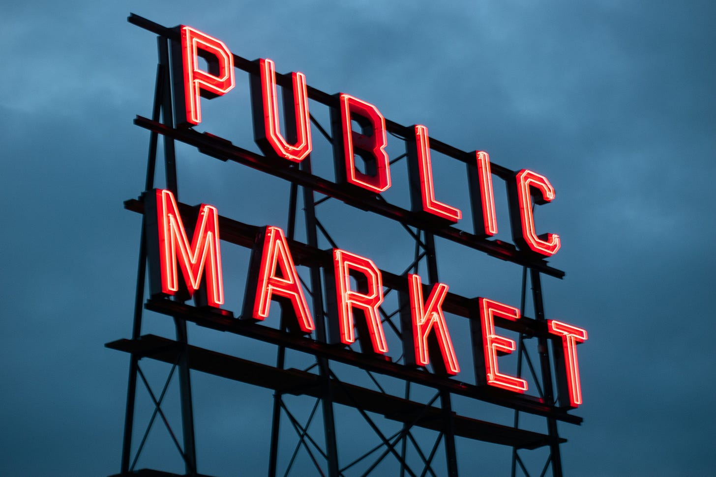 Photograph of a large neon sign that says "Public Market" positioned in front of dark clouds in the background.