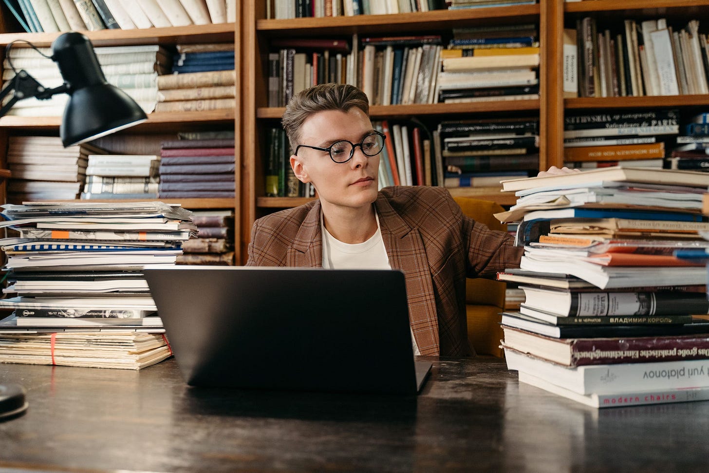 A young man with glasses working on a laptop surrounded by books.