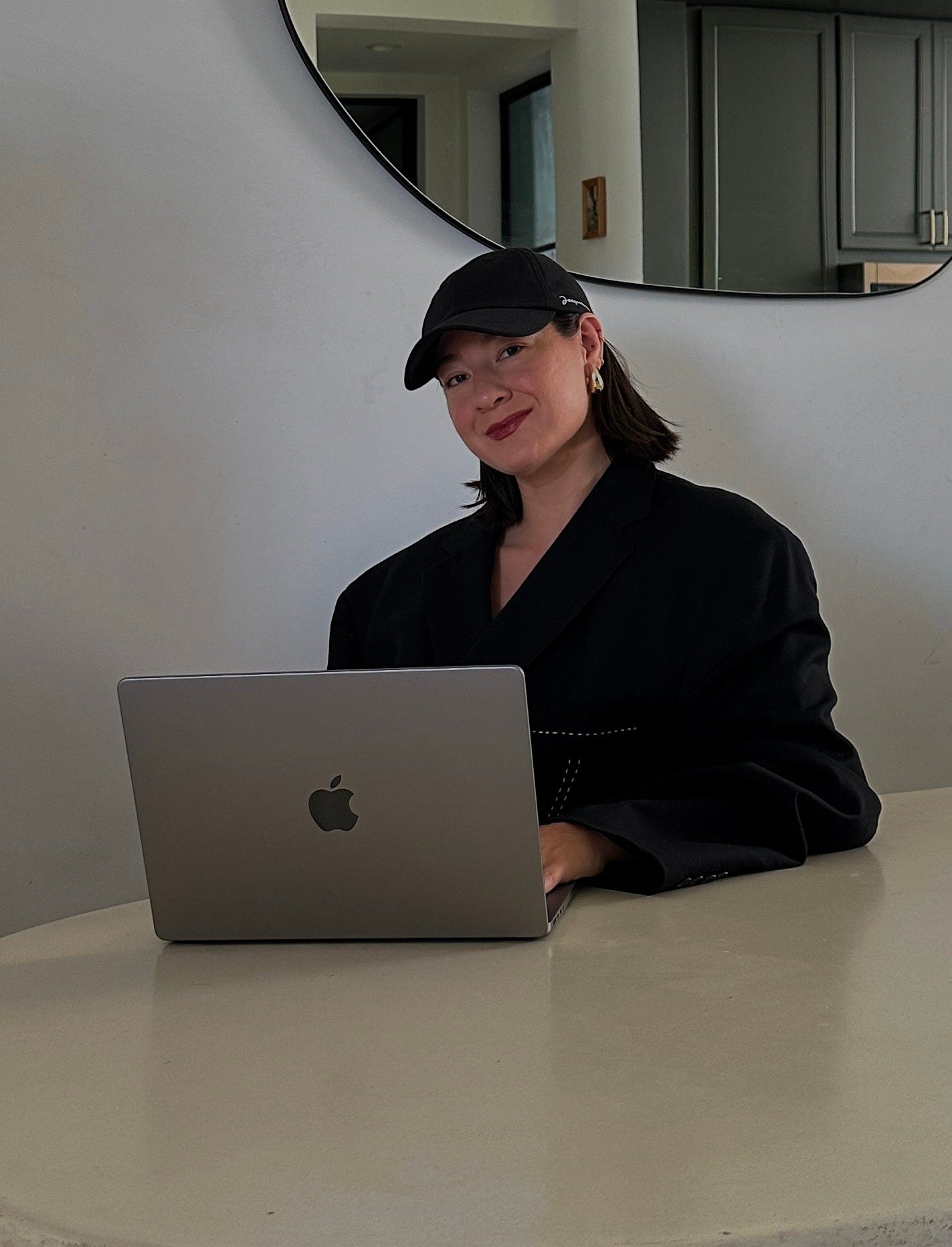 Author Melina Maria Morry sits at a white table with a mirror behind her and a MacBook while wearing a Jacquemus baseball cap and oversized black blazer: The Difference Between There, Their, and They Are