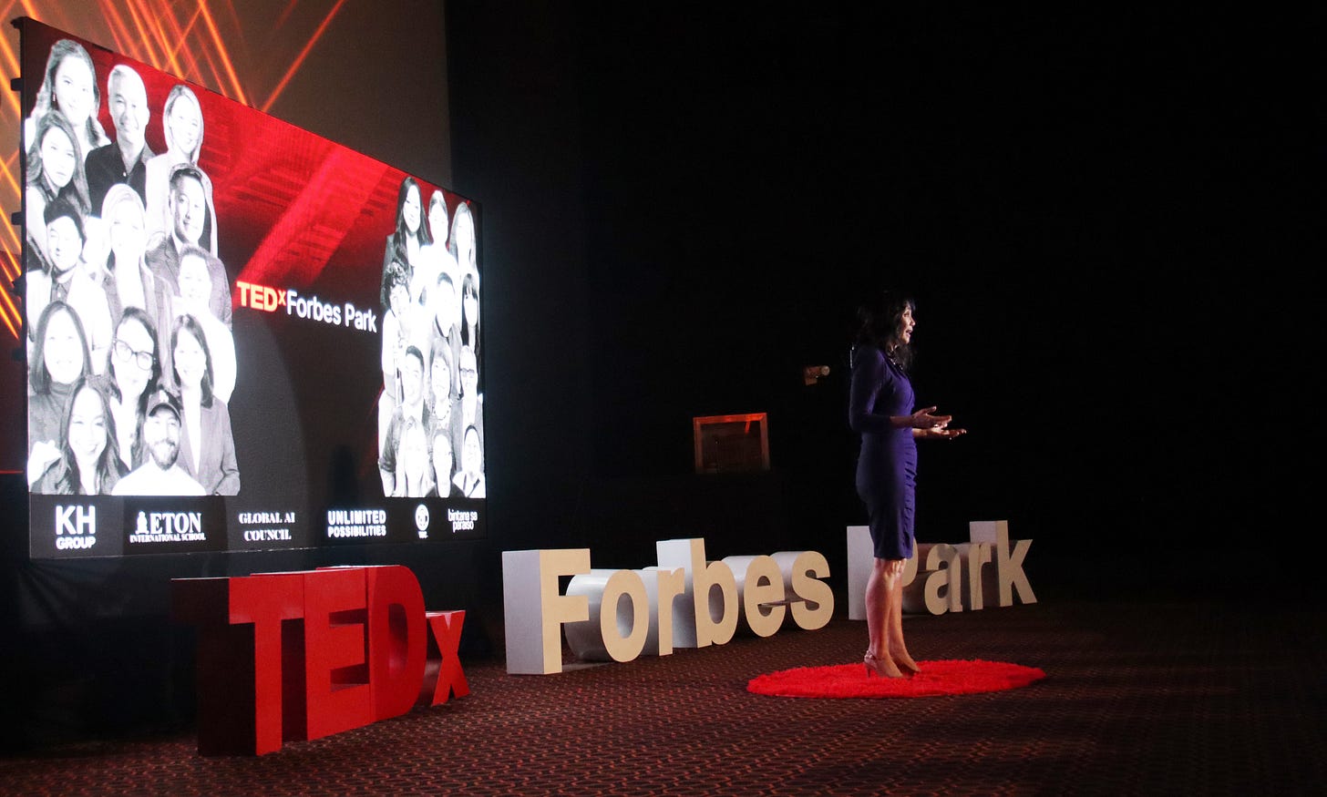 Laverne in a purple dress speaks on stage at TEDxForbesPark. Large "TEDx" letters and a red circle rug are on the floor. A screen displays black and white portraits of other speakers from that day. Laverne in a purple dress speaks on stage at TEDxForbesPark. Large "TEDx" letters and a red circle rug are on the floor. A screen displays black and white portraits of other speakers from that day.