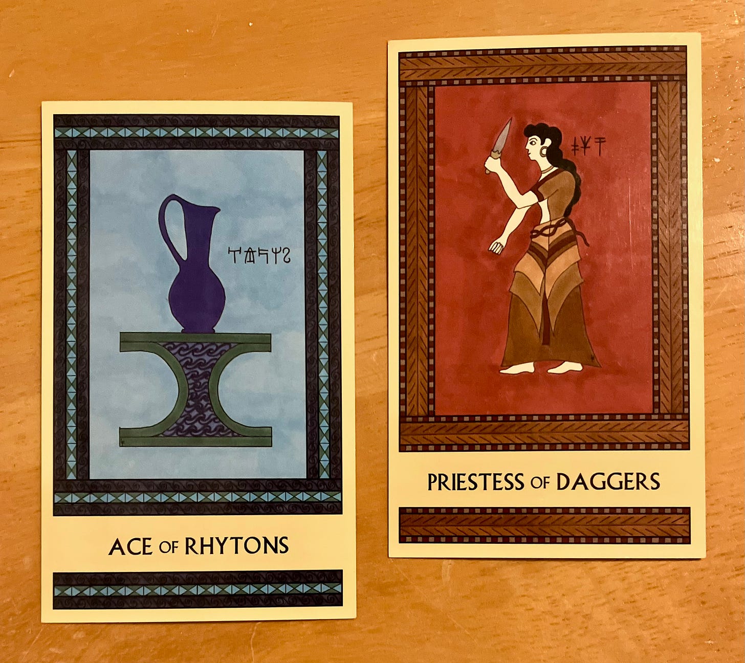 Two Minoan Tarot cards side by side on a golden brown wood surface. The Ace of Rhytons is painted in shades of blue and green. It shows a large royal blue pitcher sitting atop an incurved altar decorated with a water-wave pattern. The Priestess of Daggers is painted in shades of deep red and brown. It shows a Minoan woman standing, facing left, holding up a dagger in front of her in her left hand. 