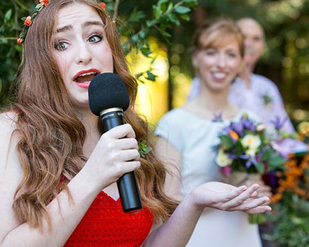 A still image of a Hannah wearing orange and holding a microphone at age fourteen, with Ilyse blurred in the background wearing white, looking at her and grinning.