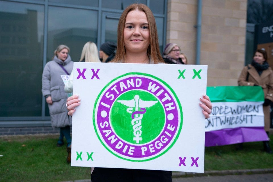 Woman holding a sign that says "I stand with Sandie Peggie".
