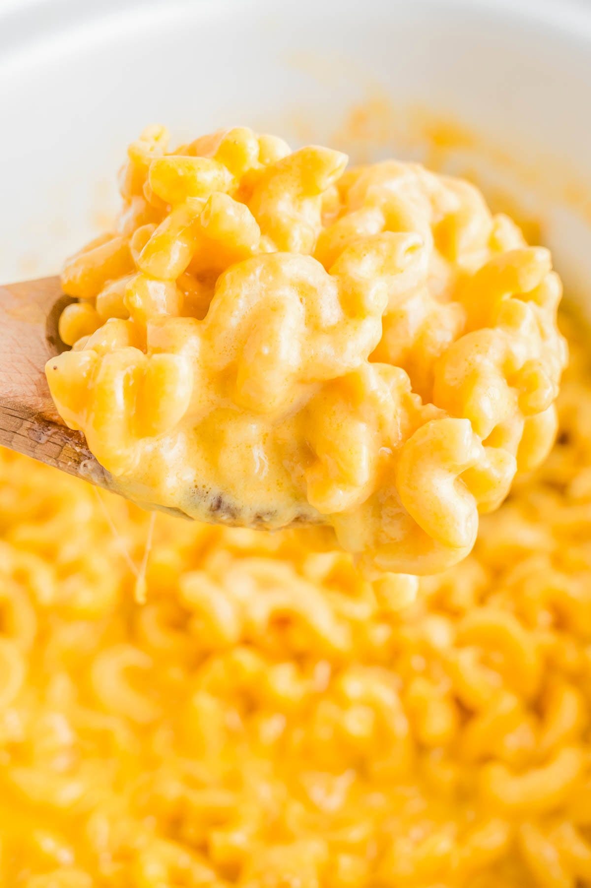 A wooden spoon holds a portion of creamy macaroni and cheese with melted cheese strings, against a background of the same dish. A wooden spoon holds a portion of creamy macaroni and cheese with melted cheese strings, against a background of the same dish.