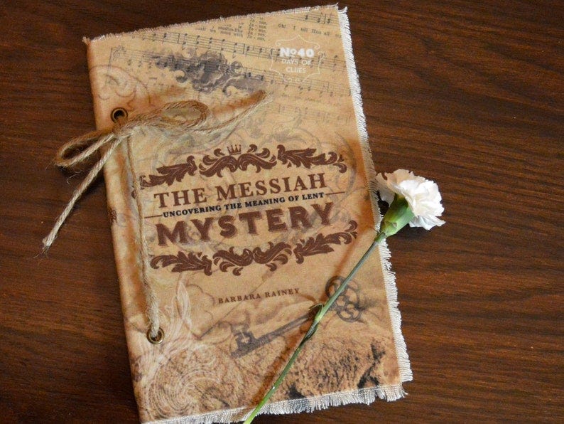 May include: A brown book with a frayed edge and a white flower on a wooden surface. The book has a title that reads 'The Messiah Mystery - Uncovering the Meaning of Lent' and the author's name 'Barbara Rainey'.