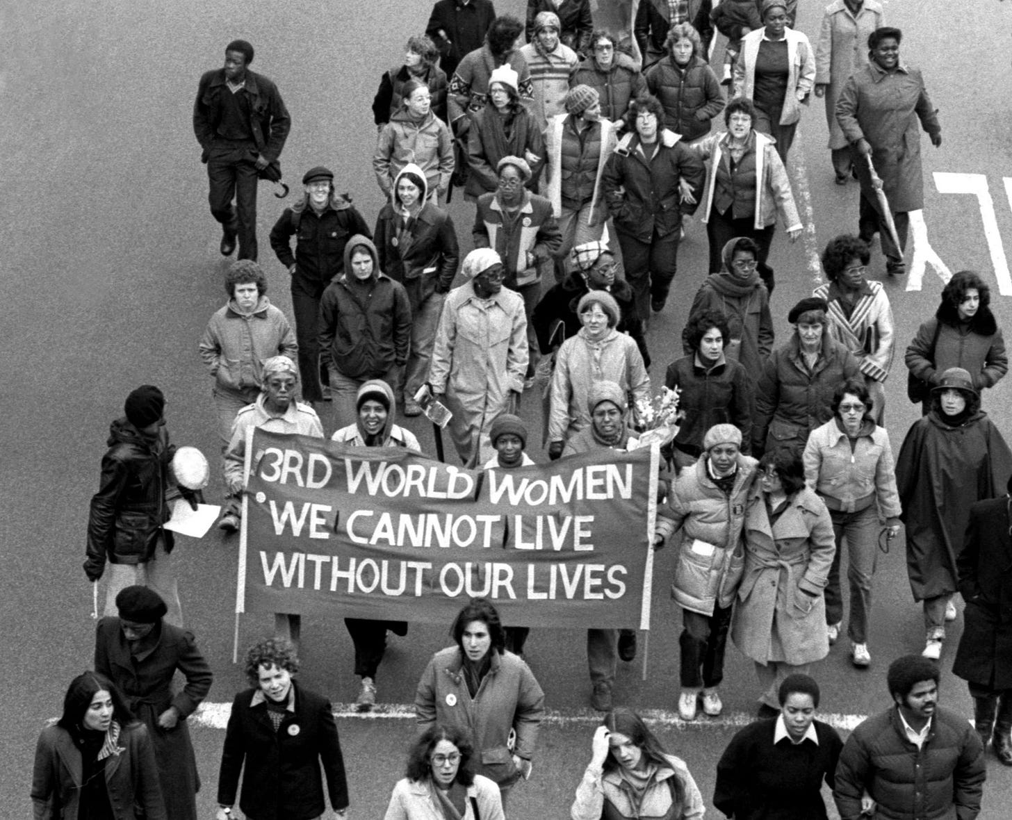 Protestors marching down a street holding a sign that reads 3rd World Women We Cannot Live Without Our Lives