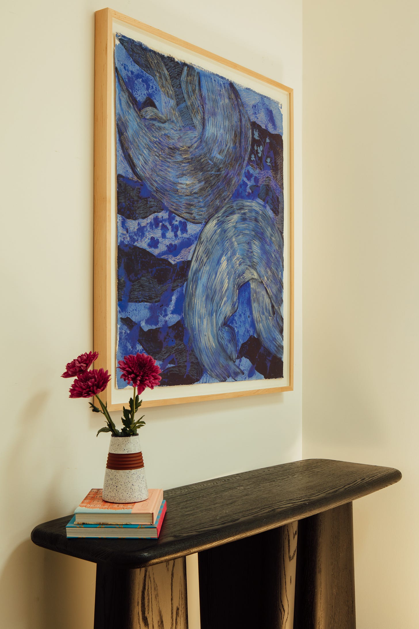 Sarana Haeata's Summer Nights, a deep blue painting of sweeping curved brushstrokes, float-mounted in a light oak frame above a blackened oak console with pink dahlias in a speckled vase on a stack of books.