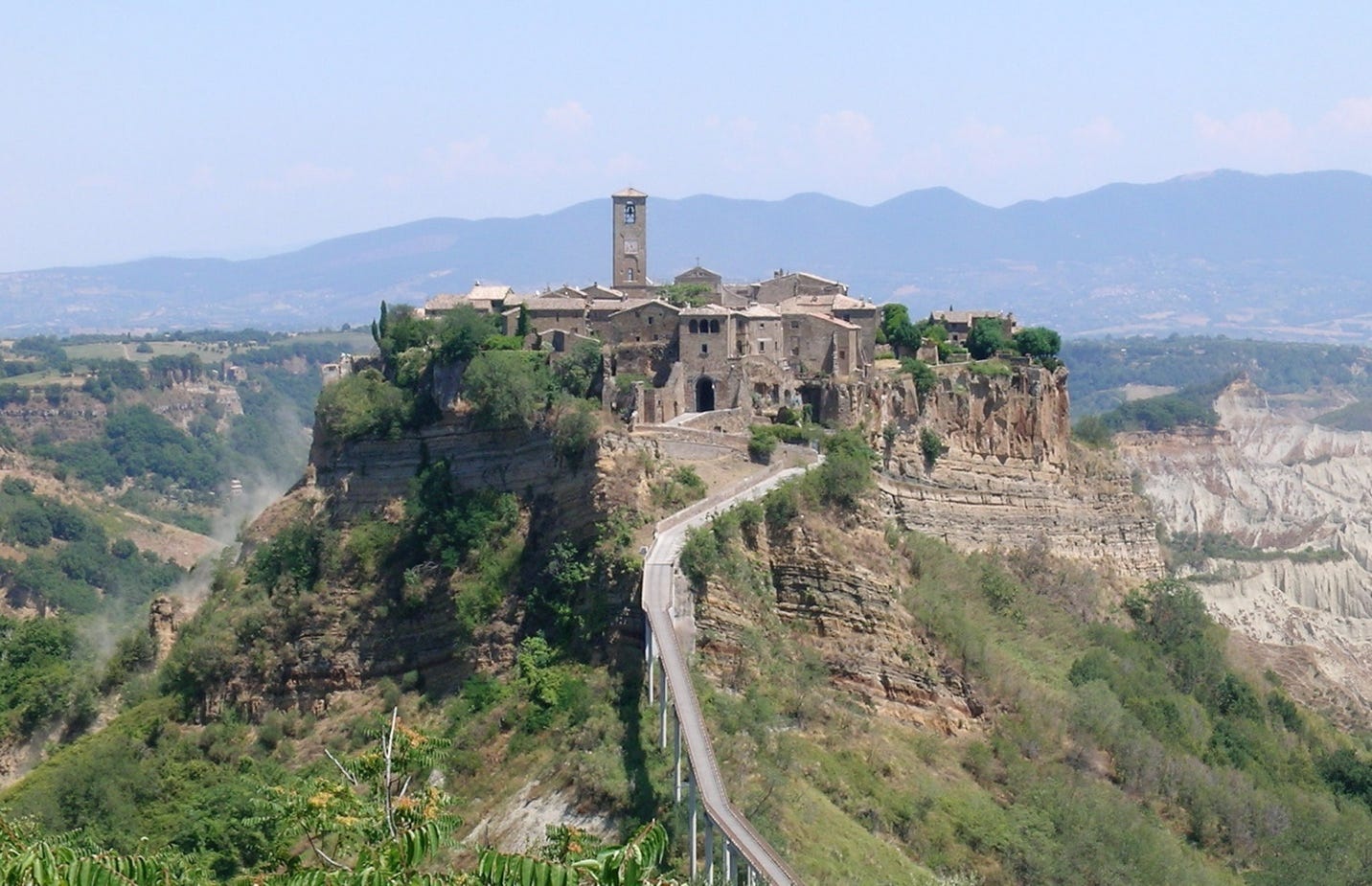 A city on a steep hill in Italy, with a road leading up to it running on a bridge. The town is clearly medieval, with a tall central tower. Civita di Bagnoregio Italy. Hill cities protected their citizens despite a huge cost in productivity. Credit Alessio Damato, CC BY-SA 3.0 <https://creativecommons.org/licenses/by-sa/3.0>, via Wikimedia Commons. Image cropped from a wide angle view by the owner.