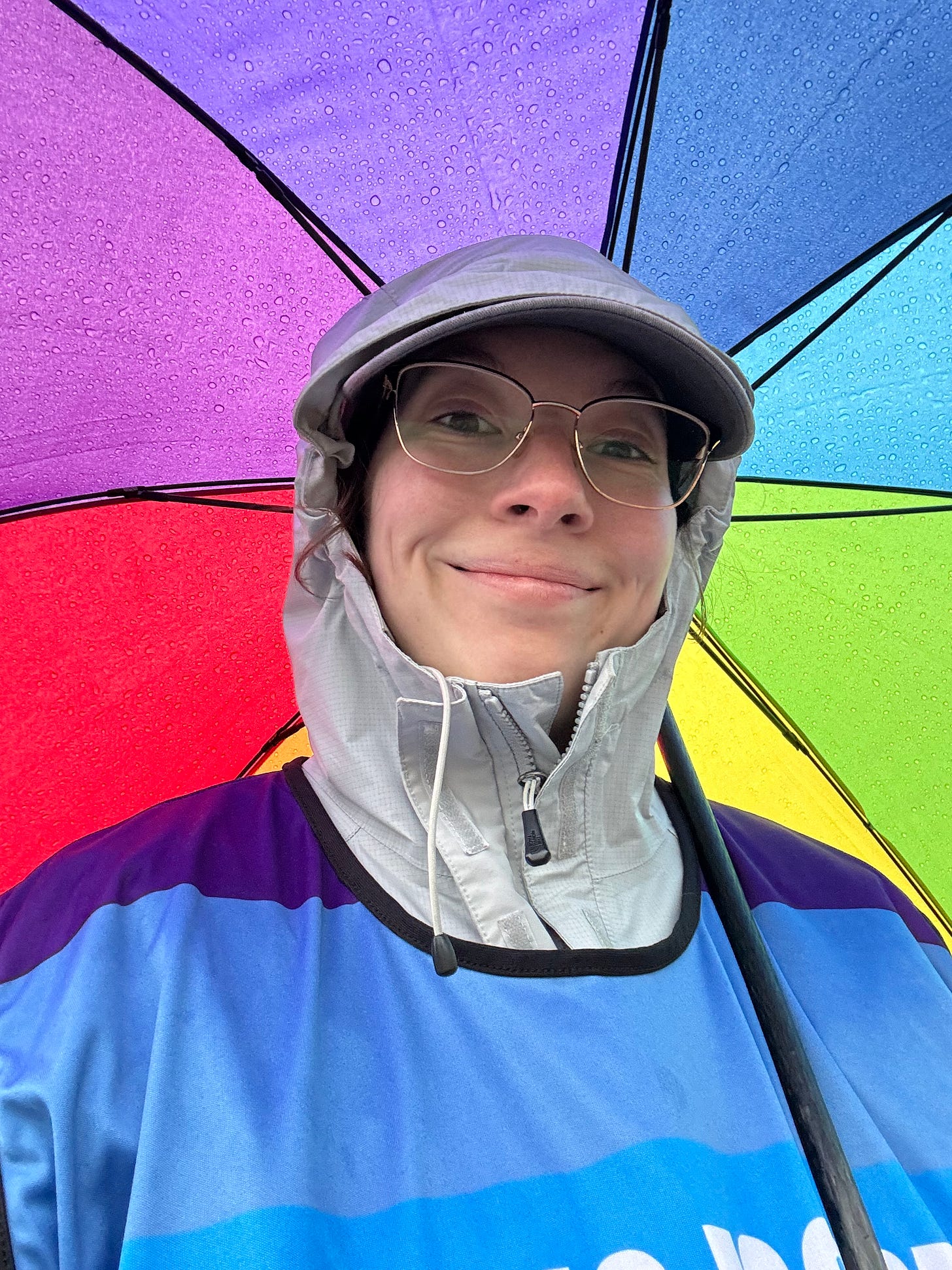 The author wears a raincoat zipped up fully, a baseball hat, and a rainbow vest. A rainbow umbrella is framed behind her