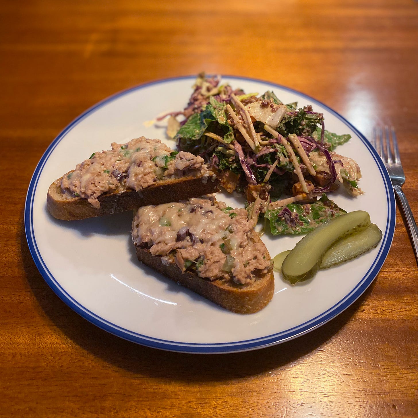 A dinner plate with an open-faced tuna melt sliced in half, pickle slices on the sad, and a pile of bright salad with yams, cabbage, greens, nuts, and apples.