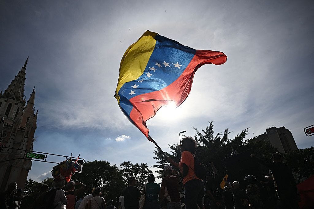 A large Venezuelan flag—yellow, blue, and red with white stars—waves dramatically in the air, backlit by the sun. The flag is held above a crowd gathered in a public square, with people seen mostly in silhouette below. Trees and buildings frame the scene, including a church spire in the background. The image conveys movement, collective presence, and a strong sense of protest or civic gathering. A large Venezuelan flag—yellow, blue, and red with white stars—waves dramatically in the air, backlit by the sun. The flag is held above a crowd gathered in a public square, with people seen mostly in silhouette below. Trees and buildings frame the scene, including a church spire in the background. The image conveys movement, collective presence, and a strong sense of protest or civic gathering.