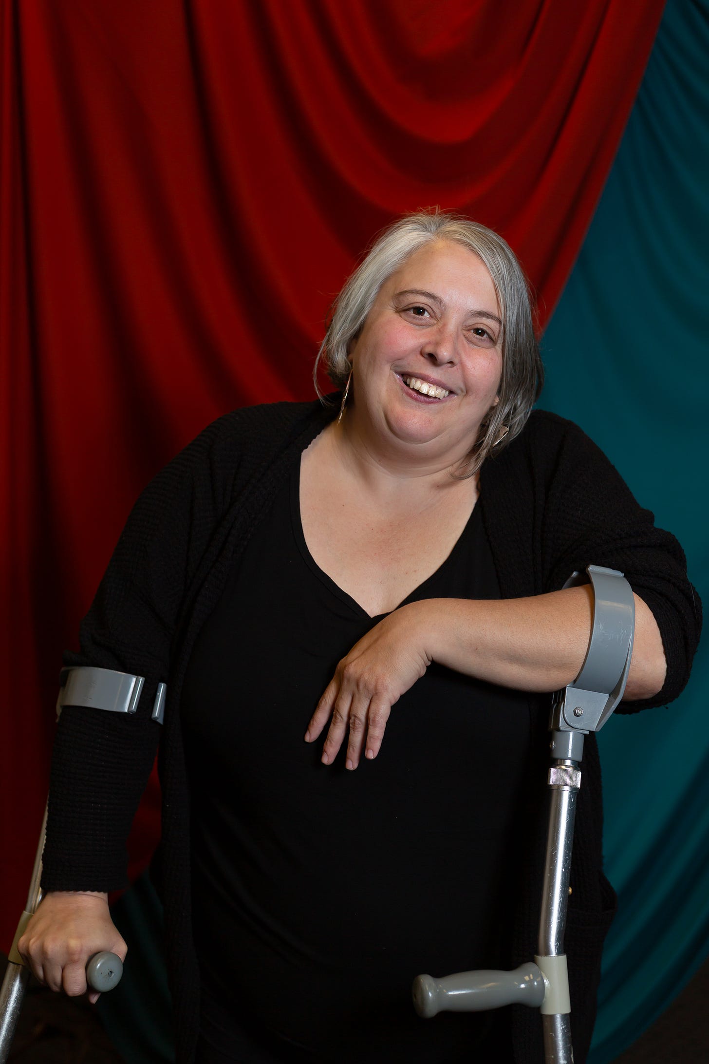 Jessica stands in front of a teal and orange backdrop. She is wearing a black top and rests on forearm crutches. She has short gray hair and silver hoops.