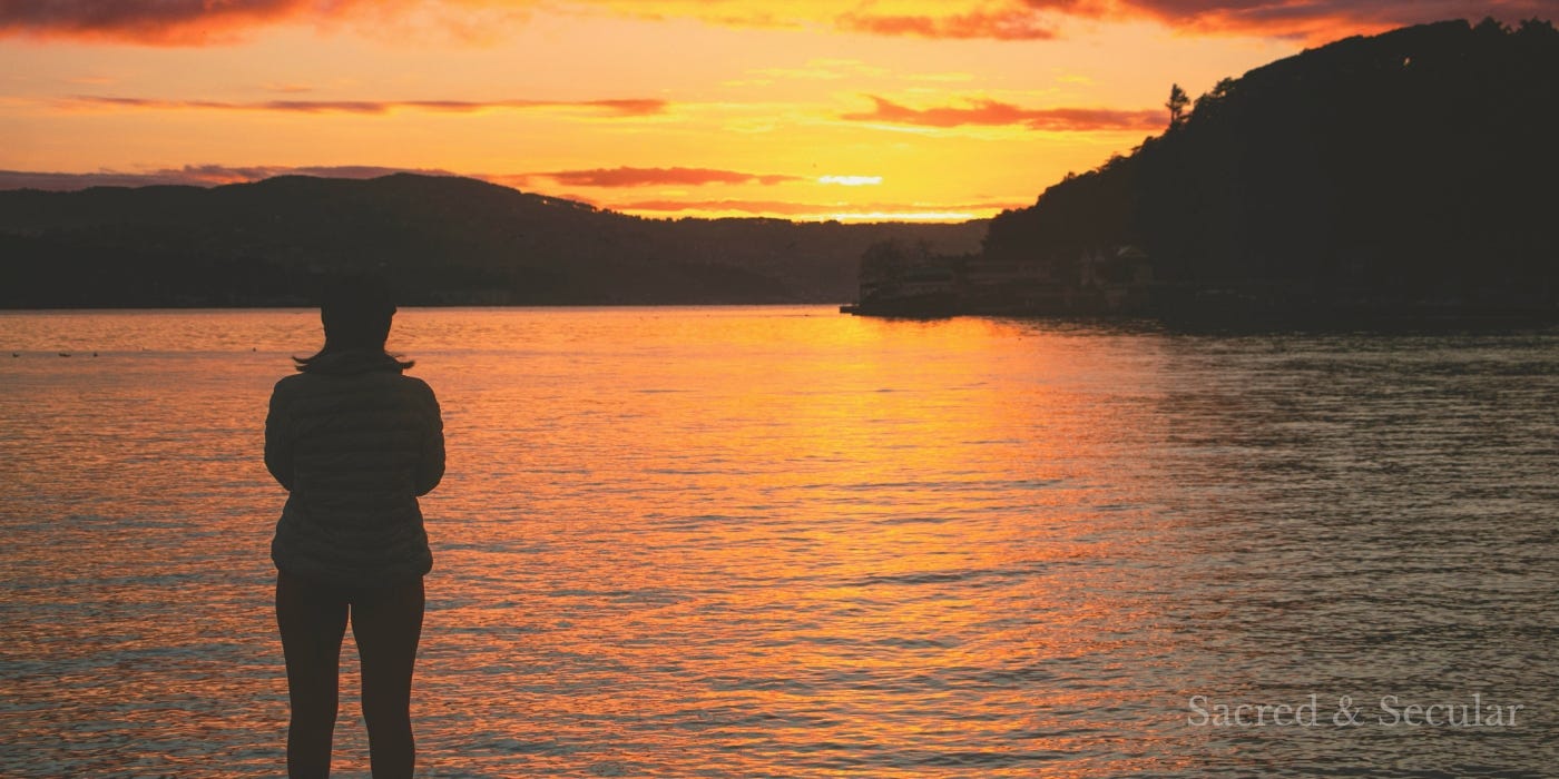 A silhouetted person standing by a calm body of water at sunset, looking out across the horizon as warm golden light reflects on the surface.