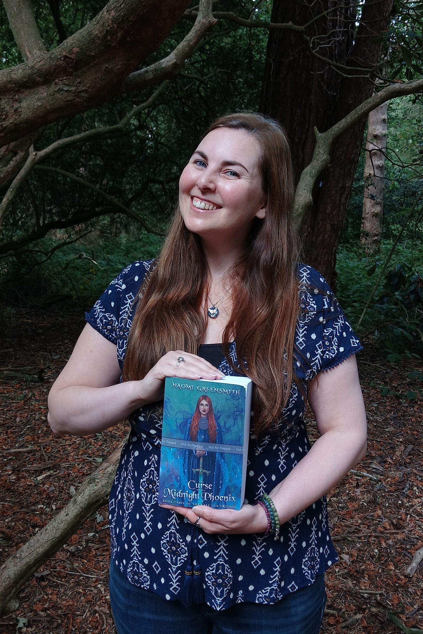 Smiling brunette woman dressed in a blue blouse and jeans standing in the forest, holding a paperback copy of The Curse of the Midnight Phoenix Smiling brunette woman dressed in a blue blouse and jeans standing in the forest, holding a paperback copy of The Curse of the Midnight Phoenix