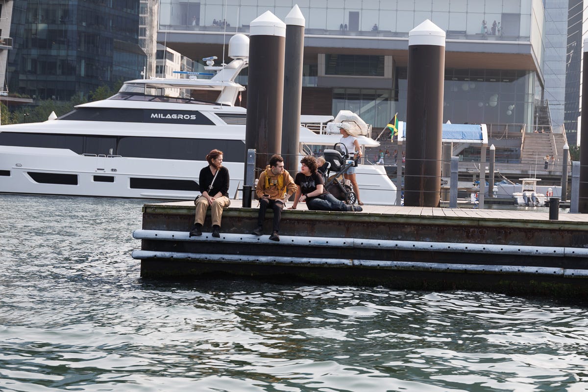 Three people sitting and talking on a dock in Boston Harbor with a large yacht in the background