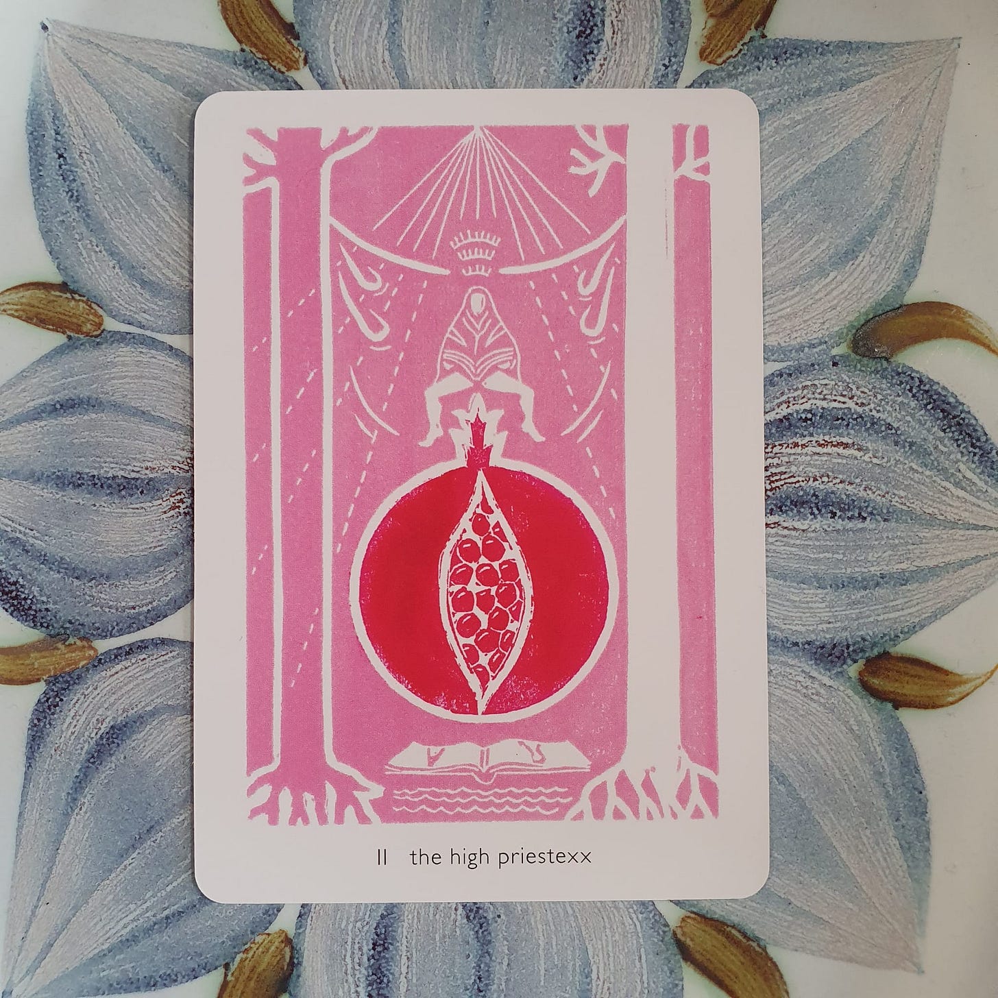 Tarot card titled “II the high priestessx” from the Queer Crow Death Magic Tarot, illustrated by Frank Duffy, showing a central red pomegranate form framed by stylised pillars, roots, and a seated figure above, in pink and red tones.