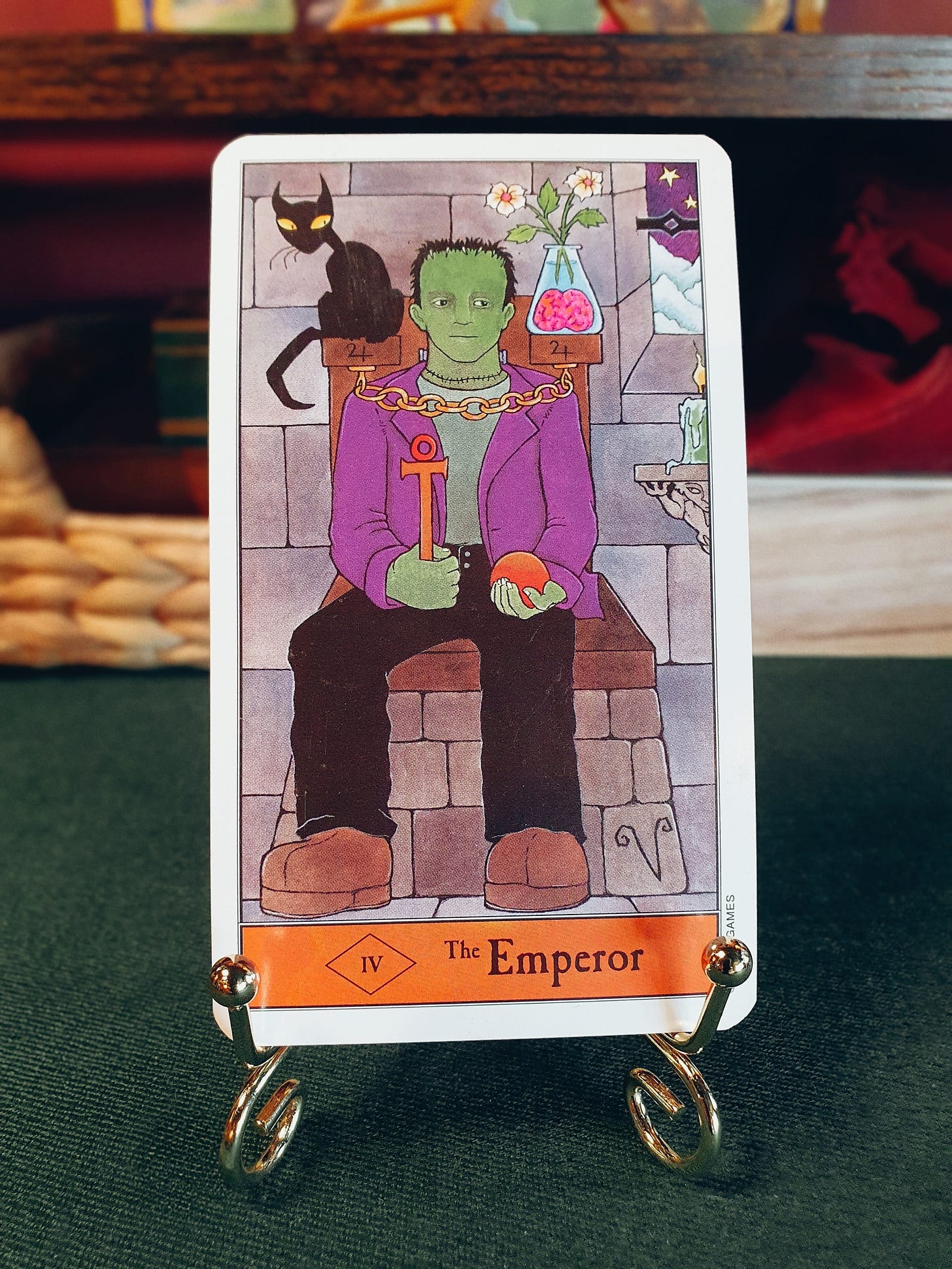 The Emperor: The Emperor tarot card from Kipling West’s Halloween Tarot, depicting a green Frankenstein-like figure seated on a stone throne, holding an ankh and an orange, with a black cat and a jar containing a brain behind him. The Emperor: The Emperor tarot card from Kipling West’s Halloween Tarot, depicting a green Frankenstein-like figure seated on a stone throne, holding an ankh and an orange, with a black cat and a jar containing a brain behind him.
