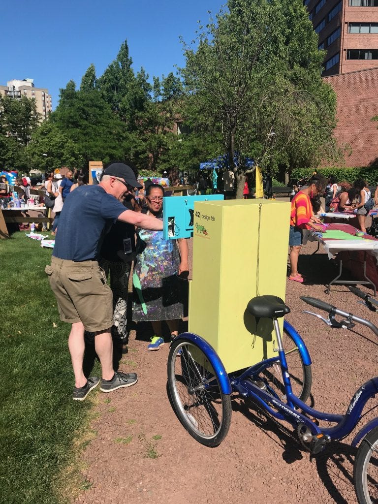 Two people are interactive with a computer screen that is mounted on a bright chartreuse green cabinet on the back of a blue adult tricycle. Two people are interactive with a computer screen that is mounted on a bright chartreuse green cabinet on the back of a blue adult tricycle.