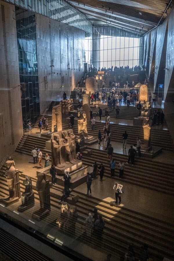 The grand, brightly lit interior of a museum hall with large stone statues of pharaohs arranged on wide, tiered staircases, surrounded by numerous visitors. The grand, brightly lit interior of a museum hall with large stone statues of pharaohs arranged on wide, tiered staircases, surrounded by numerous visitors.