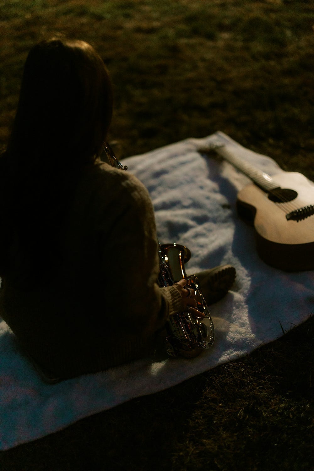 Person sitting facing away with saxophone in hand and guitar on blanket in the grass. Person sitting facing away with saxophone in hand and guitar on blanket in the grass.