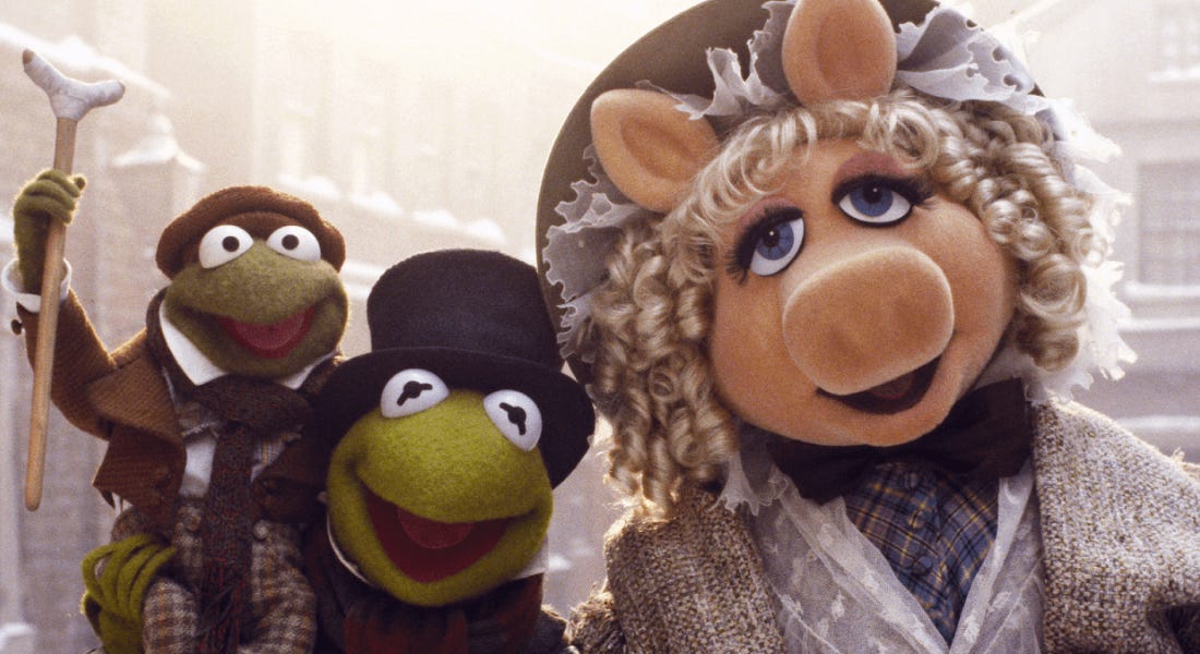 A small frog, Kermit the Frog, and Miss Piggy - three of The Muppets - wearing Victorian outfits, with a snowy town scene behind them