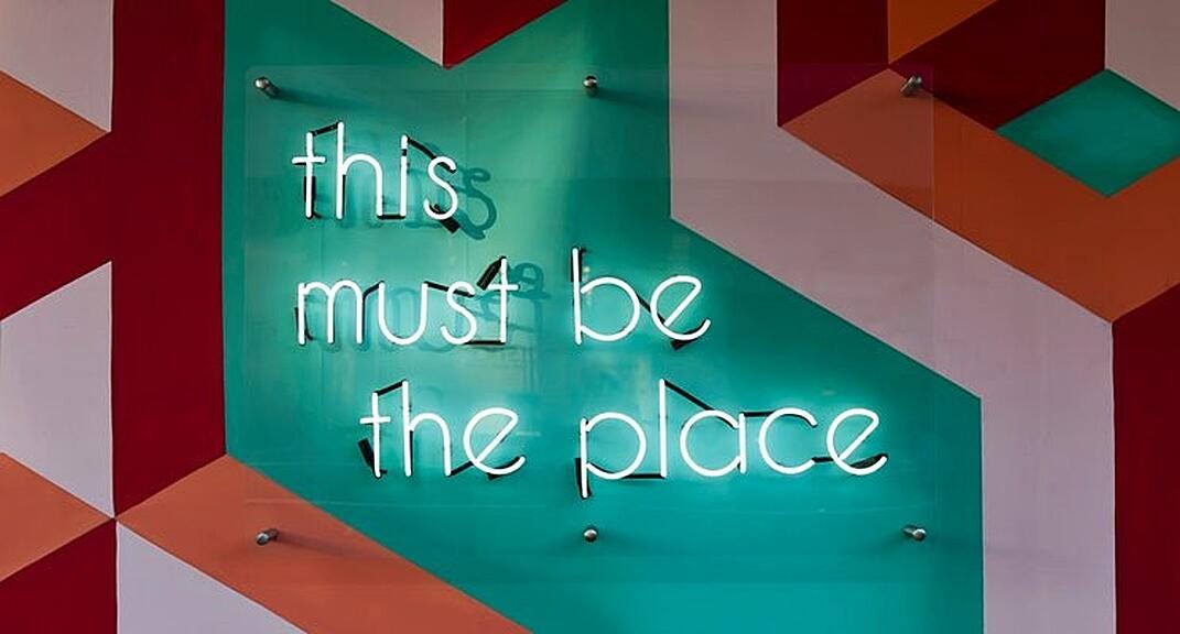 An abstract piece of art, at the centre of which is a neon sign saying ‘this must be the place.’