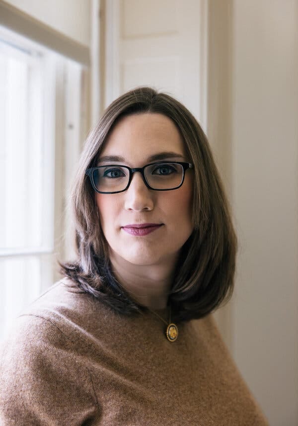 A portrait of Sarah McBride wearing a brown sweater and black glasses standing next to a window.