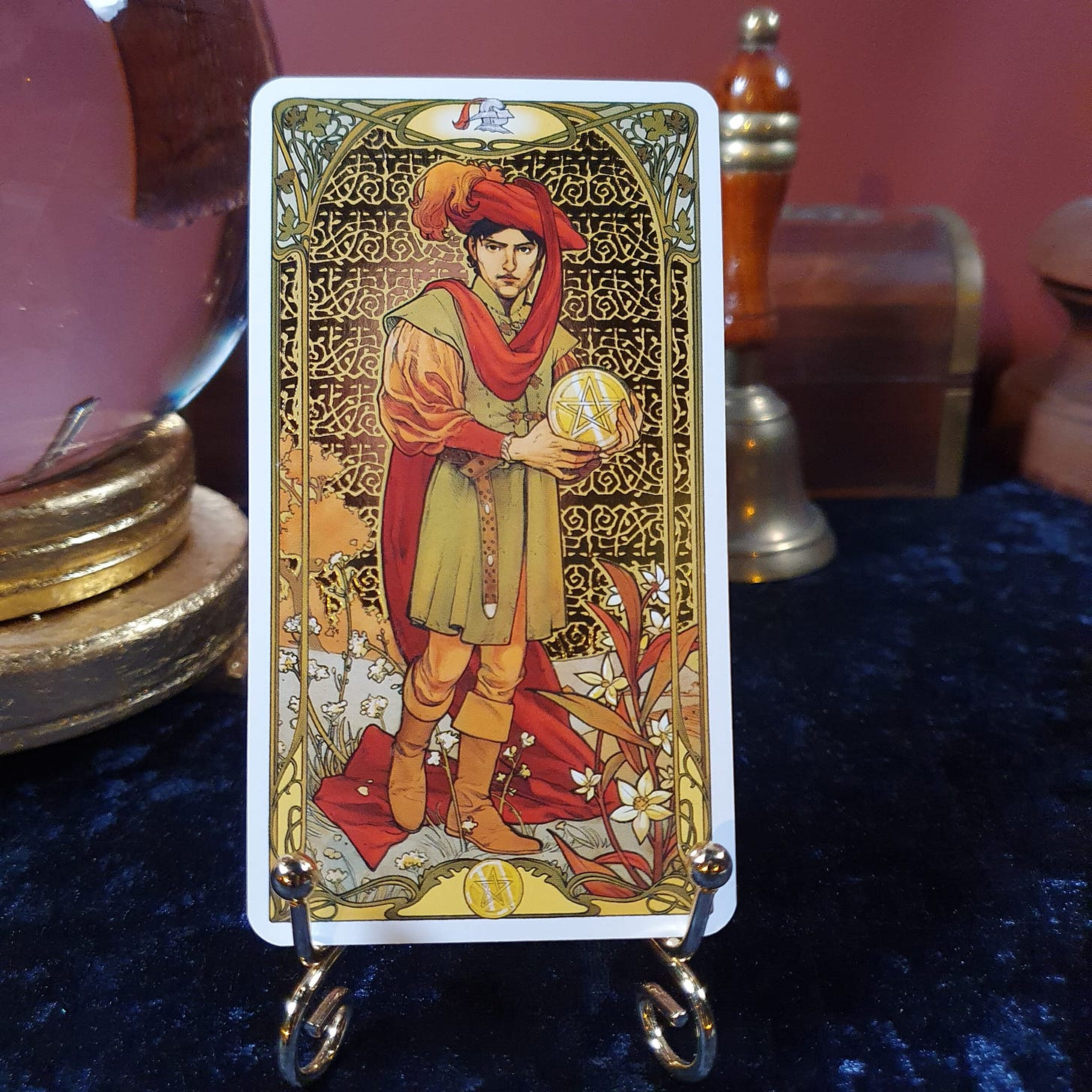 Page of Pentacles tarot card displayed on a gold stand with a crystal ball and bell in the background for the Tarot DMs interview with Elspeth Wilson.