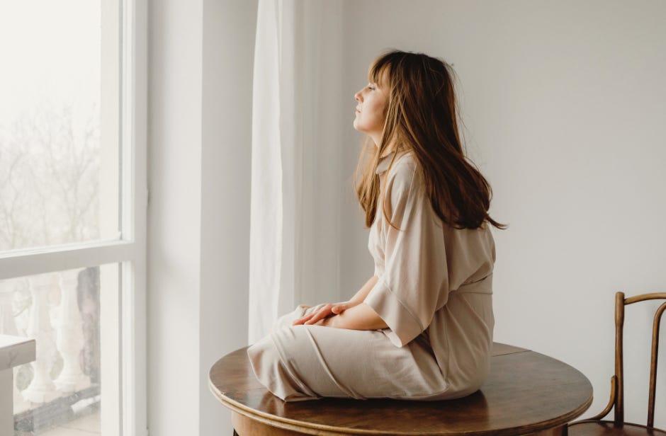 A woman sitting cross legged on a small round table, her eyes are closed and she is facing a large window with lots of daylight shining in.