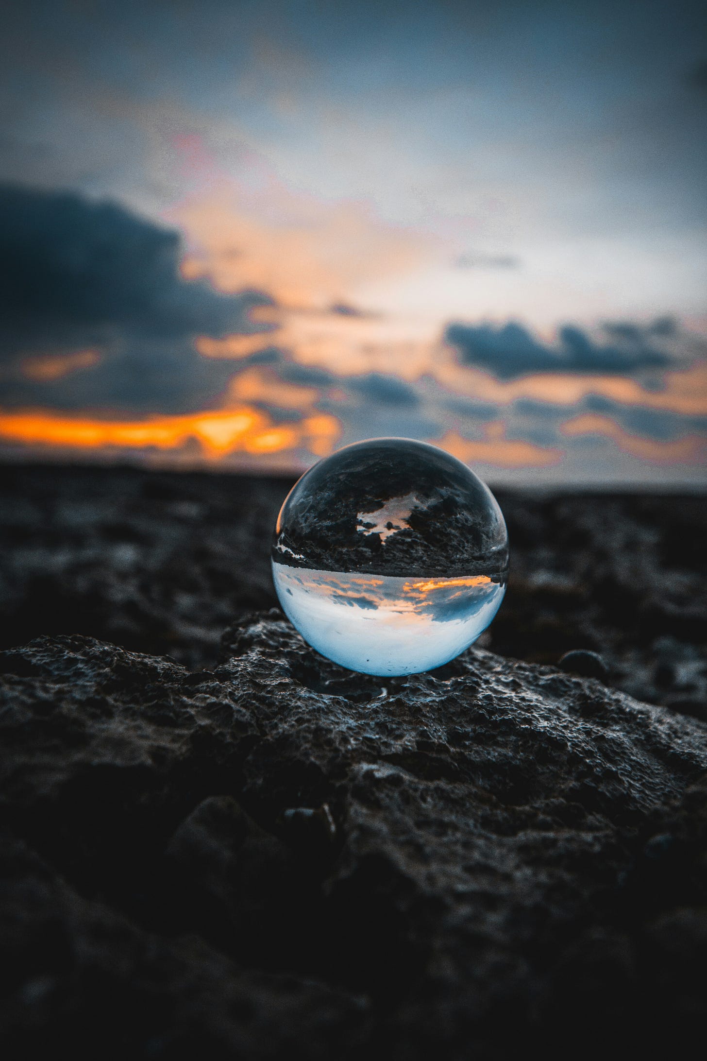 In the foreground, a clear marble rests on a dark rock. Inside the marble, the distant horizon with a sunset and clouds is flipped upside down, with the sunset on the bottom and the dark rocks and earth at the top.