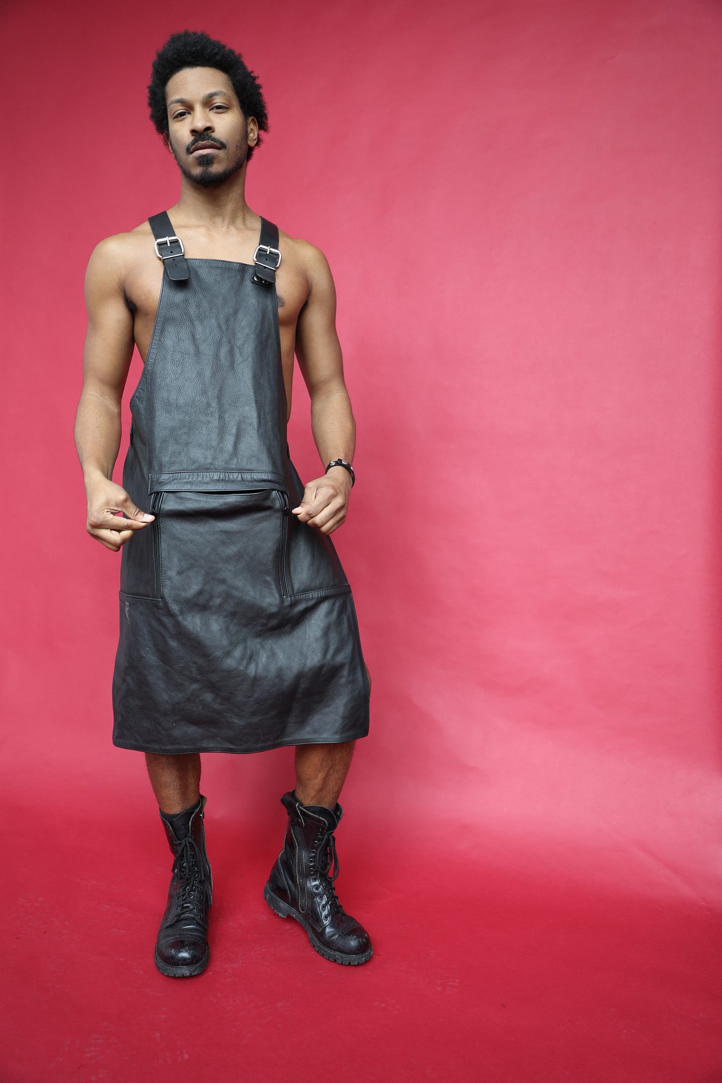 Man in black leather apron and combat boots, standing against a red backdrop, gripping the fabric at his hips. Man in black leather apron and combat boots, standing against a red backdrop, gripping the fabric at his hips.