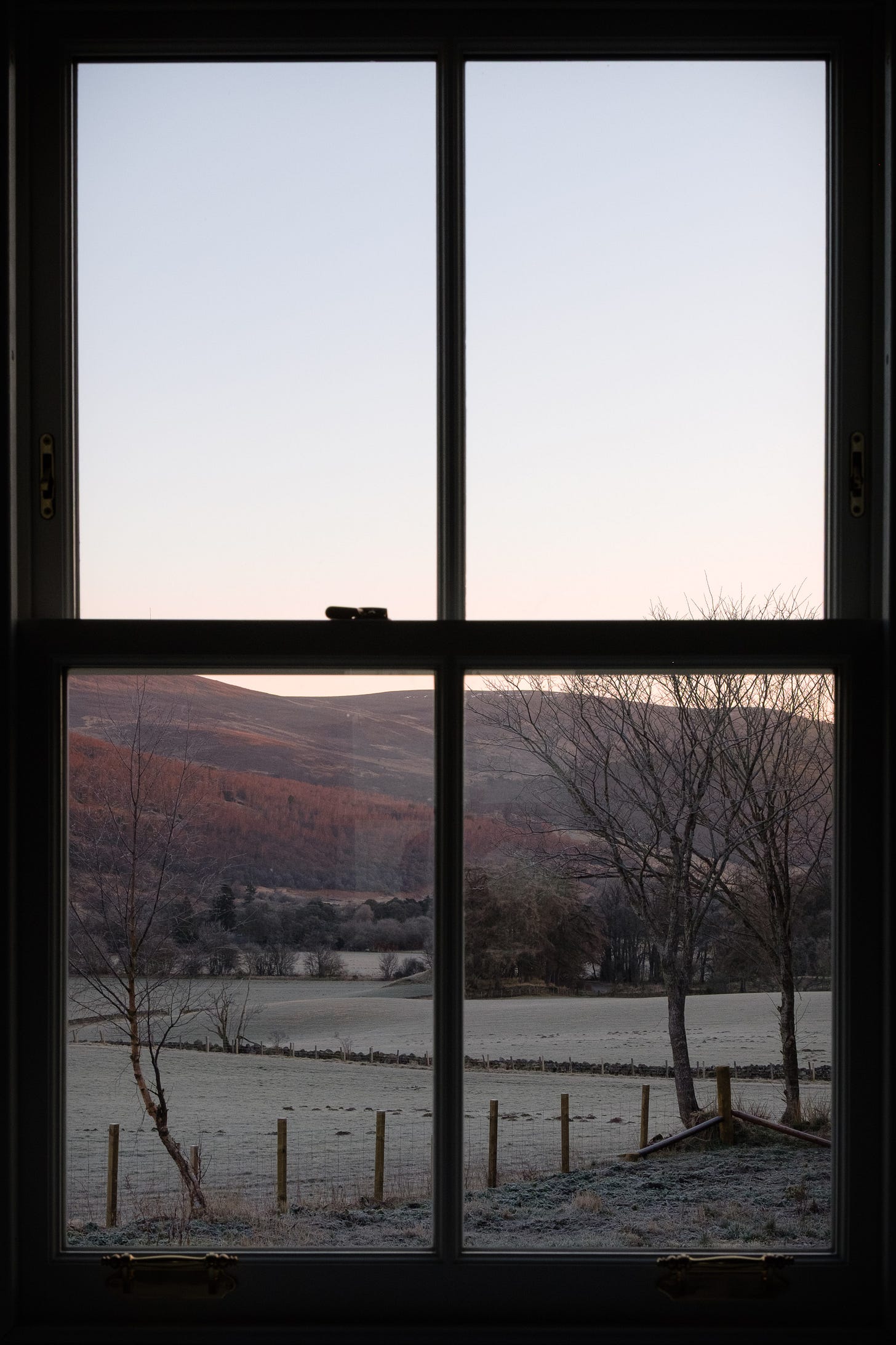 A view through a window at sunrise, showing the rolling hills of the Cairngorms dusted with frost. The sky glows with soft reds and oranges, casting a warm light across the landscape. Inside the home, the room is still and quiet, contrasting with the wide, open hills beyond. The image captures a sense of peace, transition, and the beginning of a slower life in a new place.