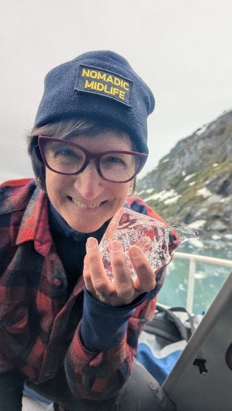  a person smiling and holding up a clear chunk of ice a little larger than her hand, with icebergs floating in the water behind her