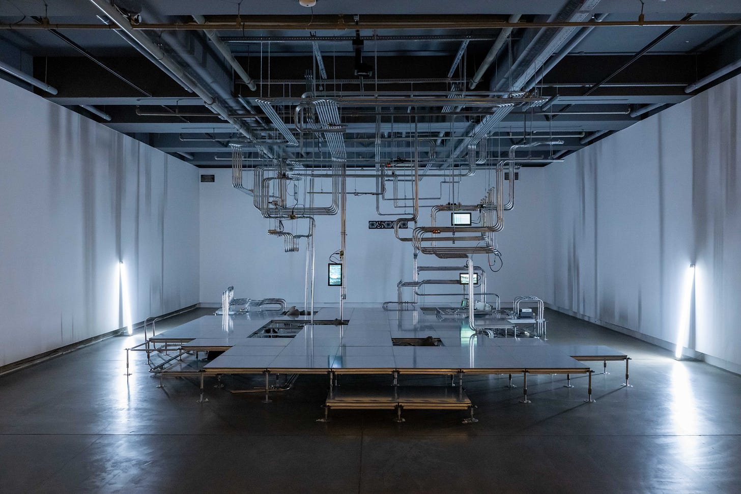 A dimly lit gallery space with spare interior fittings with a fluorescent light tube leaning against the two side walls that lights the room with an eerie blue-grey light. In the middle of the gallery, is a raised platform with a series of steel pipes, running in fixed track formation with occasional bends, that range from the ceiling to the floor in the middle of which are a number of small flat screens with graphical displays.