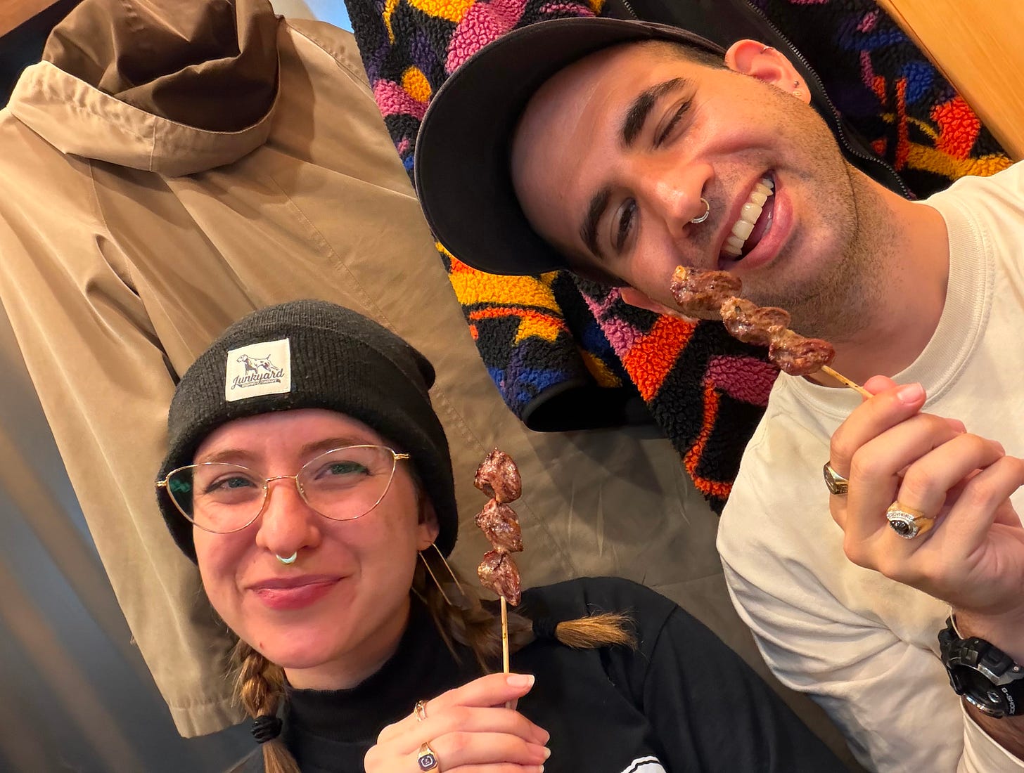 two people, one in cats eye glasses with pig tails, a beanie, brown hair, blue eyes, and a gold septum ring in a black turtleneck, the other wearing a hat with dark brown hair, a septum ring, greenish eyes, and a white long-sleeve shirt. they are sitting in front of two hanging coats, one brown, one black with a yellow, orange, pink, and purple pattern. both are holding thin wooden skewers and looking at the camera with playful faces on. the skewers each contain three cooked chicken hearts.